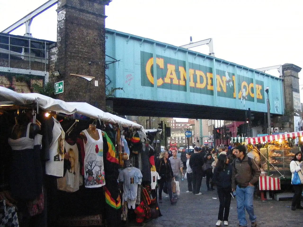Il ponte ferroviario verde acqua con la scritta Camden Lock sopra Chalk Farm Road, con bancarelle di magliette e passanti sotto