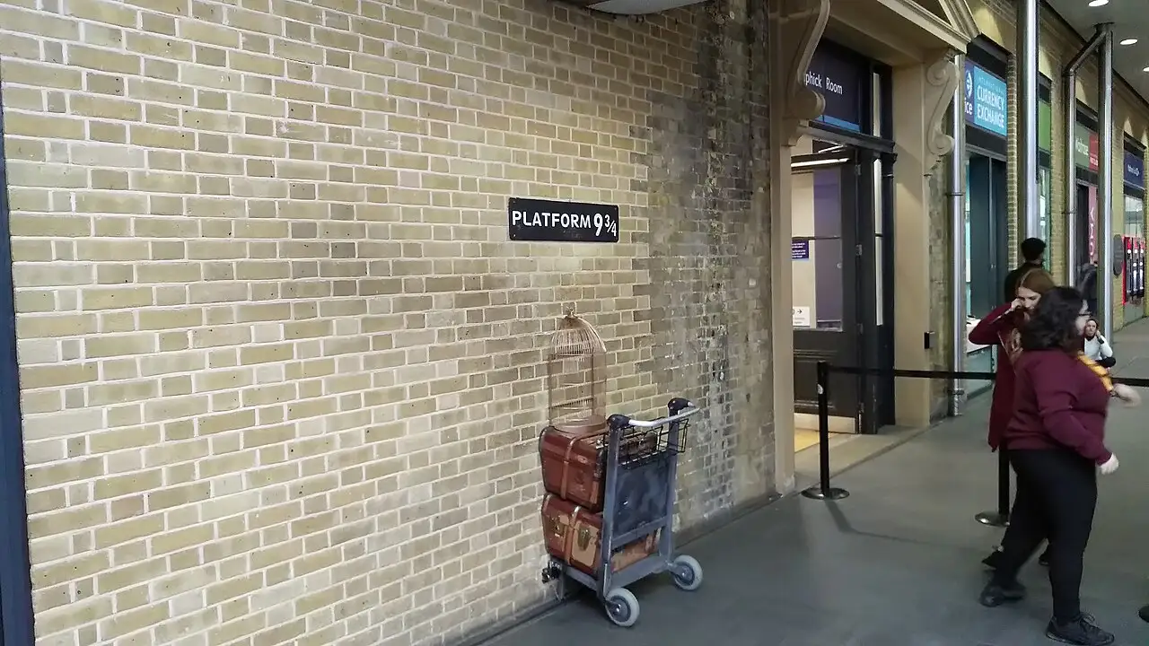 Il muro di mattoni chiari con l'insegna Platform 9¾ e un carrello con bauli e gabbia che sembra entrare nel muro, nell'atrio di King's Cross