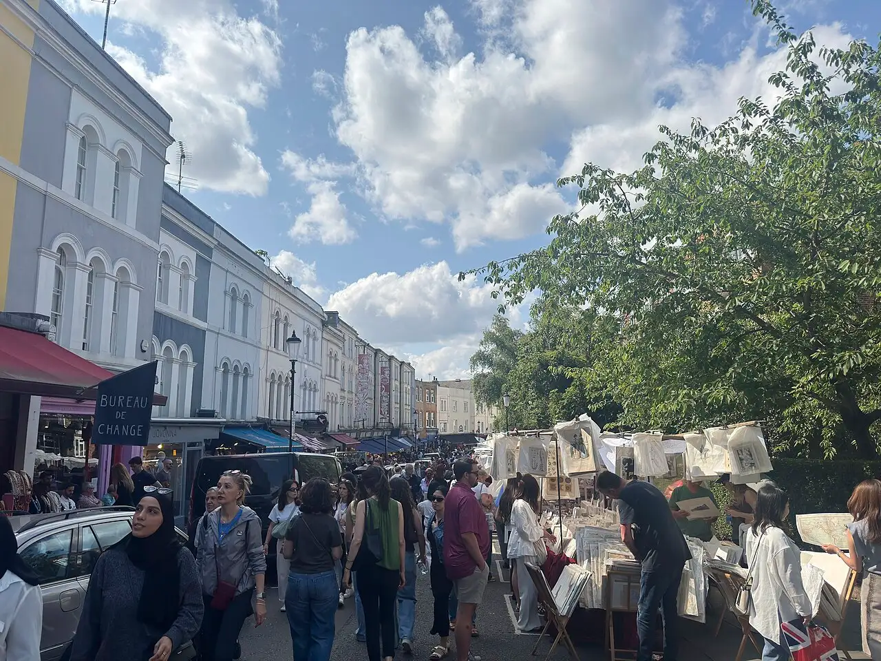 Portobello Road in un giorno di mercato: case color pastello a sinistra, banchi di stampe sotto gli alberi a destra e la folla in mezzo alla strada