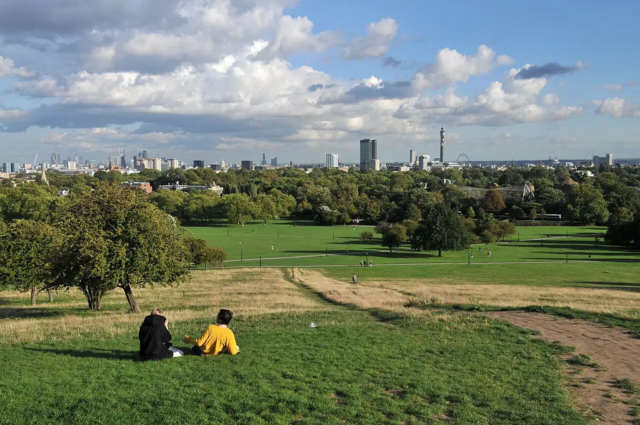 Il prato di Primrose Hill che scende verso Regent's Park, con lo skyline di Londra e la BT Tower all'orizzonte sotto un cielo di nuvole