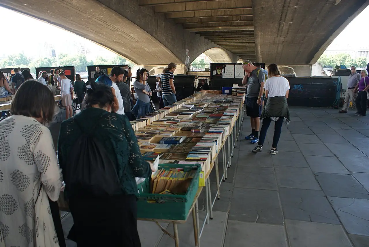 Lunghi tavoli di libri usati sotto le arcate in cemento del Waterloo Bridge, con persone che sfogliano i volumi