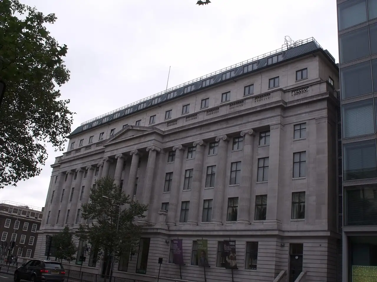 La facciata in pietra chiara del Wellcome Building su Euston Road, con le colonne ioniche e gli stendardi all'ingresso, sotto un cielo grigio